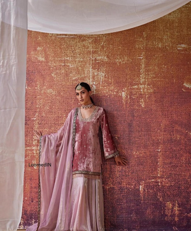 May include: A woman's ensemble featuring a dusty rose velvet tunic with long sleeves and a matching skirt. The outfit includes a light pink shawl with gold trim and a delicate beaded necklace and headpiece. The background is a textured, reddish-brown wall.