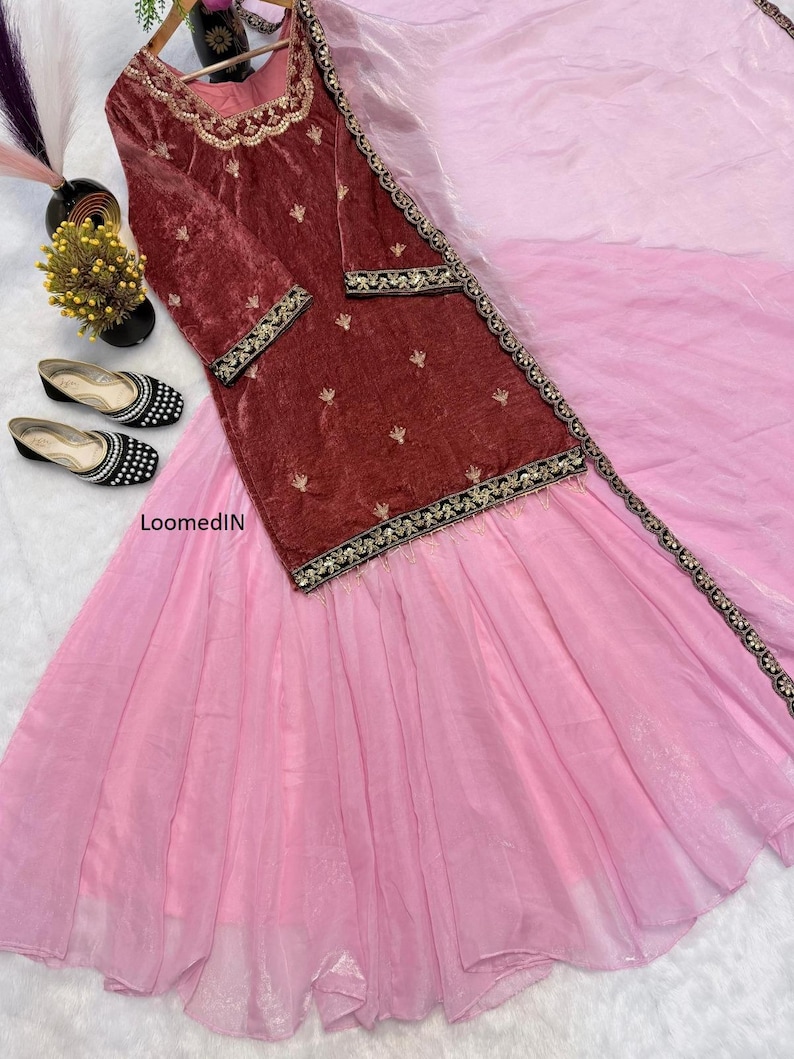 May include: A formal ensemble featuring a maroon velvet top with gold embroidery and a light pink skirt. The top has long sleeves and decorative trim. Black and white embellished flats are placed near the outfit.
