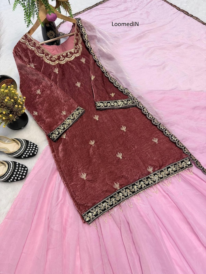May include: A maroon velvet tunic and skirt set with gold embroidery and a pink dupatta. The tunic has long sleeves and a square neckline. The skirt is a light pink color. The set is displayed on a white surface.
