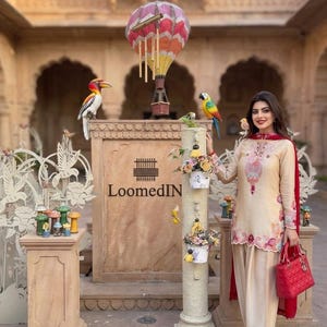 Puede incluir: Una túnica beige de manga larga con intrincados bordados florales en rosa y rojo, combinada con pantalones a juego y un dupatta rojo. El conjunto se complementa con un bolso acolchado rojo y zapatos rojos de punta fina.