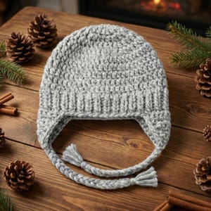 Puede incluir: Un gorro de invierno de color gris claro, tejido a ganchillo, con orejeras y lazos trenzados con borlas. El gorro se exhibe sobre una superficie de madera con piñas, palitos de canela y ramas de hoja perenne.