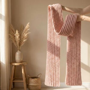 Puede incluir: Una bufanda larga de punto acanalado en color rosa suave. La bufanda se sostiene, mostrando su longitud y textura. El fondo presenta una pared de tonos neutros, una ventana con una cortina transparente y un pequeño taburete de madera con un jarrón de flores secas.