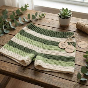 Puede incluir: Shorts de crochet con rayas crema, verde claro y verde oscuro, con cintura acanalada. Las etiquetas adjuntas dicen "Organic Yarn", "Handmade" y "Eco-Friendly". Se exhiben sobre una superficie de madera.