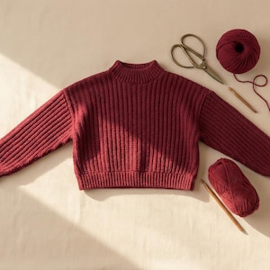 Może przedstawiać: Bordowy sweter z prążkowanej dzianiny z golfem i długimi rękawami. Na zdjęciu widać również włóczkę, nożyczki i szydełka, co sugeruje motyw rzemieślniczy. Sweter jest centralnym punktem, o miękkim, teksturowanym wyglądzie.