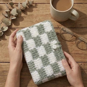 Puede incluir: Una funda de ganchillo hecha a mano con un patrón de tablero de ajedrez en verde salvia y blanco, asegurada con un cierre de botón. La funda está sostenida por manos, con una taza de café y gafas en el fondo, sugiriendo un ambiente acogedor.