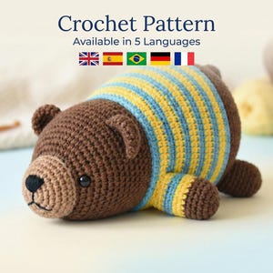 May include: A crocheted brown bear with a light brown snout and black eyes. The bear wears a blue and yellow striped shirt. The text "Crochet Pattern" is at the top of the image, with the text "Available in 5 Languages" below it.