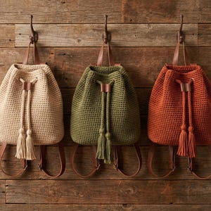 Puede incluir: Tres mochilas de ganchillo en beige, verde oliva y naranja óxido cuelgan de una pared de madera. Cada bolso tiene un cierre de cordón con borlas y correas y detalles de cuero marrón. Las bolsas están suspendidas de ganchos de metal.