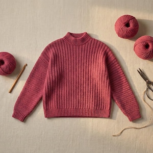 Puede incluir: Se muestra un suéter de punto acanalado de color rosa con cuello alto. Tres ovillos de hilo a juego, un ganchillo de madera y unas tijeras están colocados alrededor del suéter. Los artículos están sobre una superficie de tono neutro.