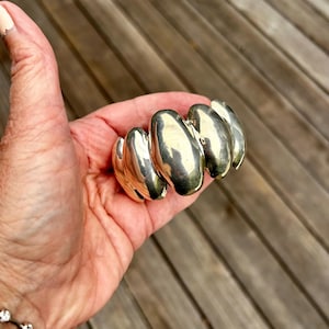 Peut inclure: Un bracelet manchette de couleur argentée composé d'une série de segments lisses, arrondis et semblables à des galets reliés entre eux. La surface métallique est hautement réfléchissante et polie, créant une forme courbe et continue.