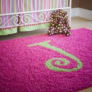 May include: A vibrant pink shag rug with a large green letter "J" design. A brown and tan polka-dot stuffed animal sits on the rug near a white crib with pink and green striped bedding.