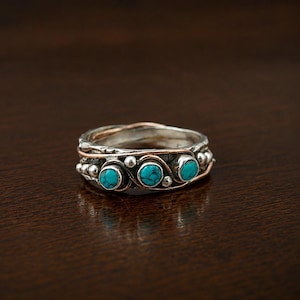 Puede incluir: Un anillo de plata con tres piedras turquesas redondas engastadas en una banda texturizada con detalles de hilo de cobre. La superficie metálica tiene un acabado oscuro y oxidado con pequeñas esferas de plata que rodean las piedras.
