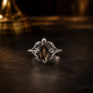 May include: A silver ring with a marquise-cut, smoky brown gemstone. The ring features a detailed leaf design on either side of the gemstone, creating an elegant and nature-inspired aesthetic. The ring is set against a dark background.