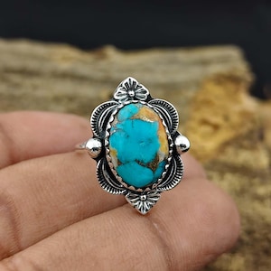 May include: A silver ring featuring an oval turquoise stone with brown matrix. The stone is set in a decorative, ornate setting with floral and beaded details. The ring is a size 7.