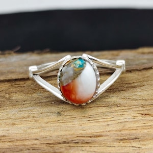May include: A silver ring with an oval-shaped stone. The stone features a mix of colors, including white, orange, turquoise, and gold. The ring's band has a split design, creating an open, airy look. The ring is displayed on a wooden surface.