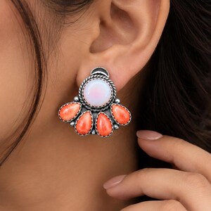 May include: A silver stud earring featuring a central round white iridescent stone surrounded by a twisted metal border, with four teardrop-shaped orange stones arranged in a fan pattern below, all set in a dark oxidised silver metal frame.