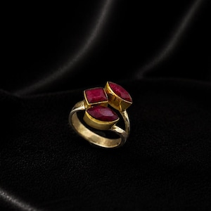 Op de afbeelding: Een gouden ring met drie grote, levendige rode edelstenen. De ring heeft een dubbelbandontwerp en de edelstenen zijn gezet in gouden bezels. De ring wordt gepresenteerd op een zwarte, getextureerde ondergrond.
