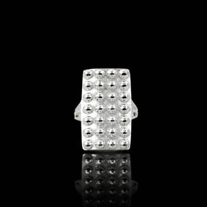 Puede incluir: Un anillo de tono plateado con una cara rectangular que presenta una cuadrícula de tachuelas esferas pulidas y elevadas. La superficie del anillo tiene un acabado mate texturizado y el diseño es simétrico, mostrando un patrón repetitivo de pequeñas protuberancias metálicas redondas.