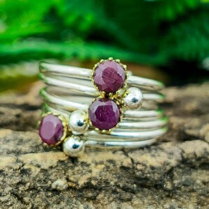 Peut inclure: Une bague en argent avec plusieurs anneaux, ornée de trois pierres précieuses rubis et de perles argentées. Les rubis sont d'un rouge profond, sertis dans des chatons dorés. La bague est présentée sur une surface brune texturée.