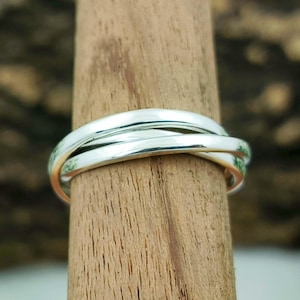 May include: A silver-colored ring with three intertwined bands. The ring is displayed on a wooden dowel, showcasing its design and shine. The bands are smooth and appear to be made of polished metal.