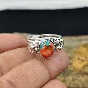 May include: A silver ring with a large, oval-shaped stone. The stone is a mix of orange, turquoise, and gold colors. The ring has a floral design on the band and small gold accents. The ring is on a person's finger.