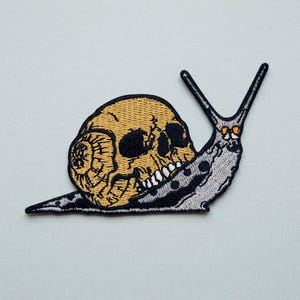 Puede incluir: Un parche bordado que muestra un caracol gris con un cráneo humano amarillo como concha. El caracol tiene dos antenas largas y pequeños ojos circulares de color naranja. Todo el diseño está delineado con hilo negro, creando una forma definida.
