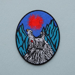 Peut inclure: Un écusson brodé ovale représentant un loup blanc avec un œil rouge hurlant vers un soleil rouge vif. L'arrière-plan montre des sommets montagneux bleus sur un ciel bleu foncé, le tout entouré d'une épaisse bordure noire brodée sur le tissu.