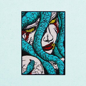 Puede incluir: Un parche bordado rectangular que muestra un rostro estilizado parcialmente oculto por gruesos tentáculos texturizados de color azul verdoso. El diseño incluye ojos amarillos, detalles rojos alrededor de los ojos y labios, y un pequeño anillo dorado.