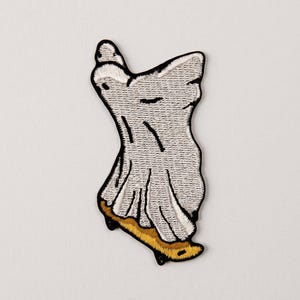 Könnte beinhalten: Ein gestickter Aufnäher, der eine weiße Geisterfigur in einem Laken auf einem gelben Skateboard zeigt. Der Geist hat schwarze gestickte Umrisse und Details, wobei das gesamte Motiv von einem dicken schwarzen Fadenrand umgeben ist.