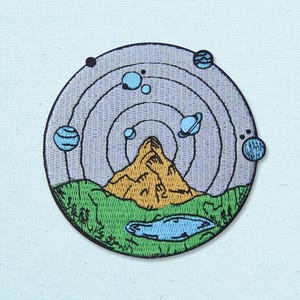 May include: A circular embroidered patch featuring a landscape with a golden mountain and a blue lake in the foreground. The sky is light blue with concentric circular lines and several small blue planets orbiting the mountain peak.