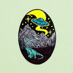 May include: An oval embroidered patch featuring a teal long-necked dinosaur standing in a dark landscape with grey mountains. Above, a bright yellow crescent moon illuminates a blue UFO emitting a beam of light, surrounded by small yellow stars.
