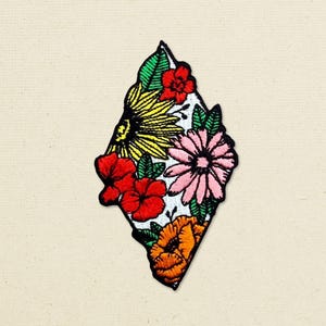 May include: A diamond-shaped embroidered patch featuring a vibrant floral arrangement. The design includes a large yellow sunflower, a pink daisy, bright red hibiscus-style flowers, and an orange blossom, all accented with green leaves.