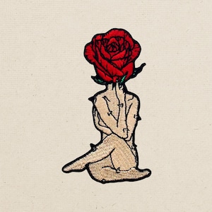 Puede incluir: Un parche bordado que muestra una figura sentada con una gran rosa roja como cabeza. La figura tiene un tono de piel beige claro, está representada en posición sentada con las piernas cruzadas y rodeada de pequeños acentos negros en forma de espinas.