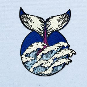 May include: An embroidered patch featuring a whale tail emerging from a circular blue background filled with stylized white ocean waves. The design uses dark blue, light blue, white, and purple thread to create a detailed marine scene.