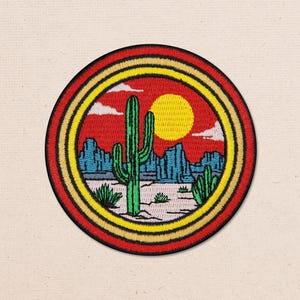 Może przedstawiać: Okrągła haftowana naszywka przedstawiająca pustynny krajobraz z zielonym kaktusem saguaro, niebieskimi formacjami skalnymi i jasnym żółtym słońcem na tle czerwonego nieba. Wzór jest otoczony koncentrycznymi pierścieniami w odcieniach żółci, pomarańczu i czerwieni.