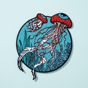 Peut inclure: Un écusson brodé circulaire représentant deux méduses aux chapeaux rouges et aux yeux bleus, flottant sur un fond bleu clair avec des motifs d'algues et de coraux bleu foncé. Les méduses ont de longs tentacules blancs et rouges.