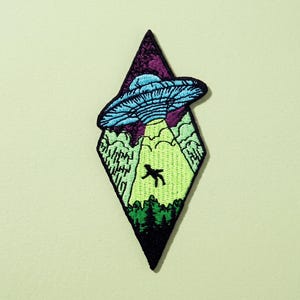 Op de afbeelding: Een ruitvormige geborduurde patch met een lichtblauwe UFO boven een donkergroen bos. Een felle geelgroene lichtstraal komt uit het vaartuig en verlicht het zwarte silhouet van een persoon die wordt ontvoerd.