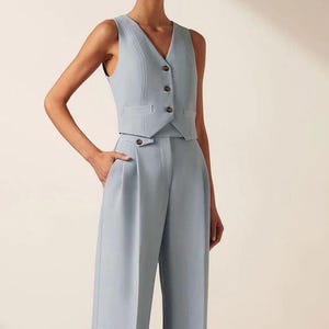 Puede incluir: Un chaleco sin mangas de color azul claro con cuello en V y tres botones oscuros, combinado con pantalones a juego de talle alto y pernera ancha. Los pantalones presentan un detalle de botón en el lateral de la cintura y una silueta entallada.