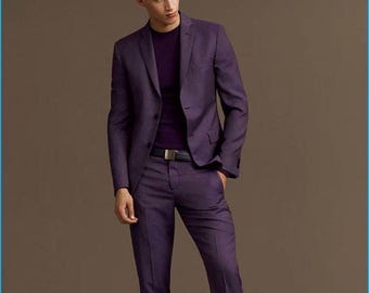 May include: A man in a purple suit, comprising a blazer and trousers, with a purple sweater. The outfit is completed with a black belt and black dress shoes. The background is a plain brown.