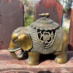 Puede incluir: Figura de elefante de color bronce con detalles intrincados. El elefante tiene una silla decorativa con un recorte floral y un pequeño remate en la parte superior. La figura se exhibe sobre una superficie de madera.