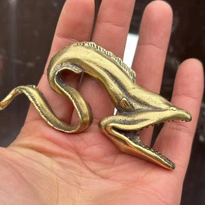 Può includere: Statuetta di anguilla in ottone con bocca aperta e squame dettagliate. L'anguilla è arricciata in una forma sinuosa, con una superficie strutturata e una tonalità dorata. La statuetta è tenuta in una mano.