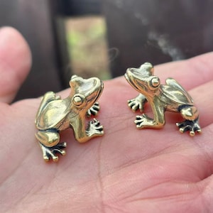 Puede incluir: Dos pequeñas figuras de rana de latón. Las ranas están detalladas con ojos grandes y patas palmeadas. Las figuras están posadas como si se estuvieran mirando. El latón tiene un color dorado cálido.