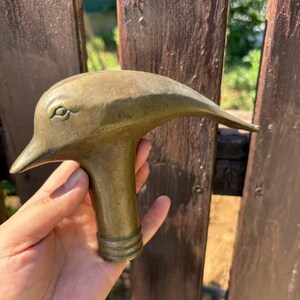 May include: A brass cane handle shaped like a dolphin head with a smooth, metallic finish. The handle features a cylindrical base with three horizontal engraved rings near the bottom, designed to attach to a walking stick or cane shaft.