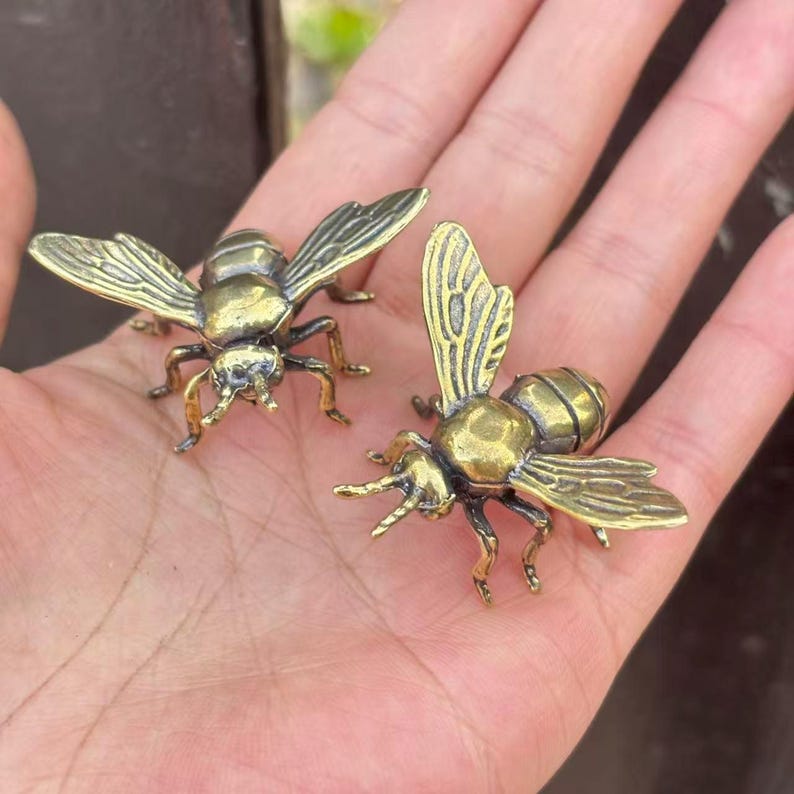 K&ouml;nnte beinhalten: Zwei kleine, detaillierte Bienenfiguren aus Messing mit ausgebreiteten Fl&uuml;geln. Die Bienen sind goldbraun mit komplizierten Fl&uuml;gelmustern und zarten Beinen. Die Figuren werden in einer Hand gehalten.