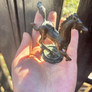 May include: Bronze horse figurine with a detailed mane and tail, standing on a circular base with embossed details. The horse is posed mid-stride, showcasing its muscular build. The figurine has a weathered appearance.