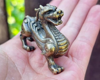 Puede incluir: Pequeña figura de criatura mítica de color bronce, posiblemente un dragón o una bestia similar. La figura está detallada con escamas, una melena y una expresión feroz. La criatura está en posición de pie, con las patas delanteras extendidas. El fondo está borroso.