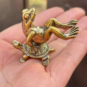 May include: A small, detailed brass figurine depicting a frog riding on the back of a turtle. The frog is in an upright position, and the turtle has an ornate shell design. The figurine has a polished, antique finish.