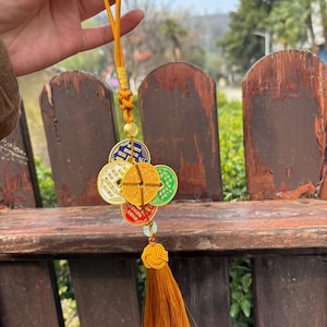 May include: A gold-colored charm featuring five circular coins in various colors, including red, green, and yellow. The charm is attached to a yellow cord with a tassel. The coins have Chinese characters on them.