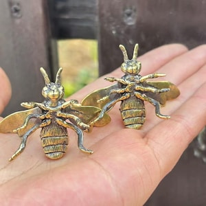 K&ouml;nnte beinhalten: Zwei detaillierte Bienenfiguren aus Messing mit ausgebreiteten Fl&uuml;geln und aufwendigen K&ouml;rperdetails. Die Bienen, in Gold- und Silbert&ouml;nen, werden auf einer Hand pr&auml;sentiert. Diese dekorativen Artikel sind ideal f&uuml;r die Inneneinrichtung.
