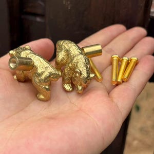 May include: Two gold-colored bear-shaped cabinet knobs with textured surfaces and protruding threaded metal posts, accompanied by three separate gold-colored threaded screws for installation, all displayed in a palm.