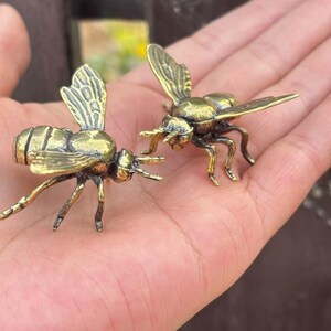 K&ouml;nnte beinhalten: Zwei kleine, detaillierte Messing-Bienenfiguren. Jede Biene hat komplizierte Fl&uuml;gel- und Beindetails mit goldgelbem Finish. Die Bienen sind so positioniert, als w&uuml;rden sie interagieren und ihre F&uuml;hler und Stacheln zeigen.